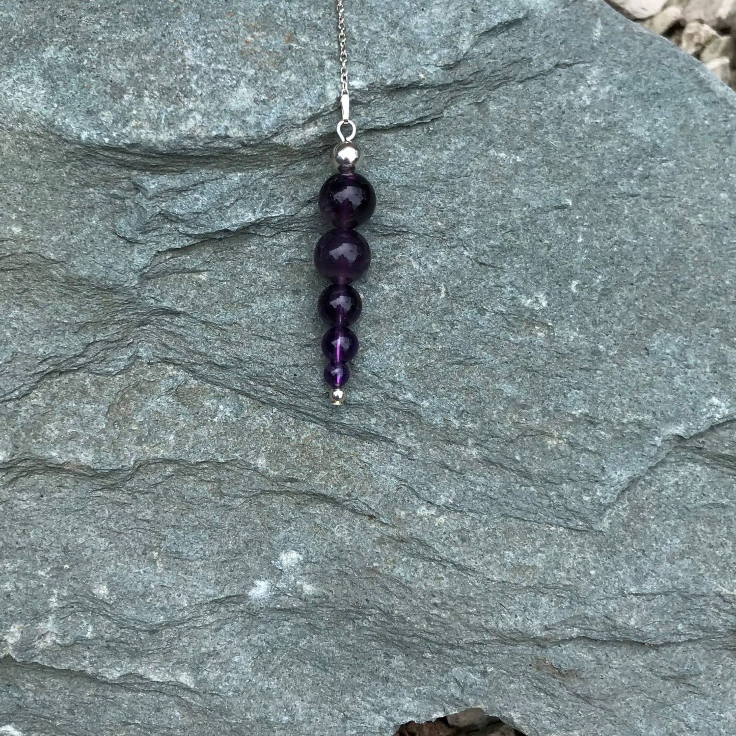 A beaded amethsyt pendulum outside on a stone.